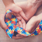Mãos de um adulto e de uma criança segurando um laço colorido de peças de quebra-cabeça, símbolo de conscientização do autismo.