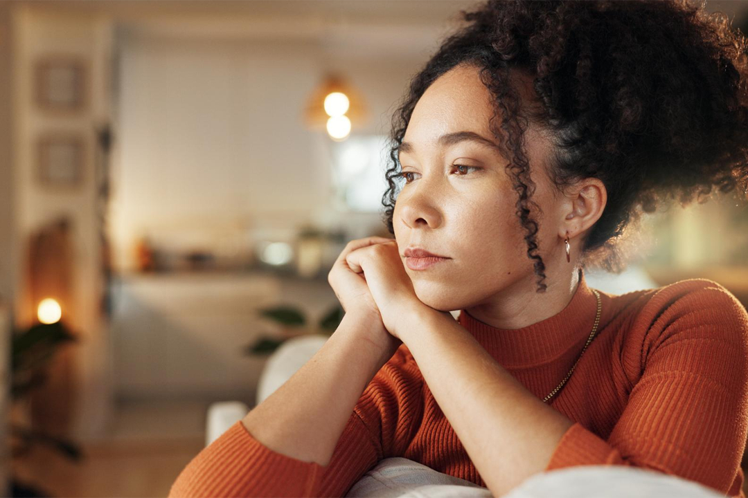 Mulher sentada em casa, com expressão pensativa, apoia o rosto nas mãos em um ambiente acolhedor e silencioso.