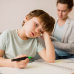 Criança sentada à mesa durante as férias, desenhando com expressão de cansaço e distração, enquanto um adulto observa ao fundo, em ambiente doméstico.