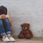 Criança encolhida no canto ao lado de urso de pelúcia retrata a ansiedade infantil autista.