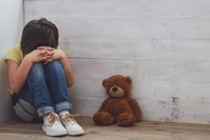Saiba mais sobre ansiedade infantil autista com a salz! Criança encolhida no canto ao lado de urso de pelúcia retrata a ansiedade infantil autista.