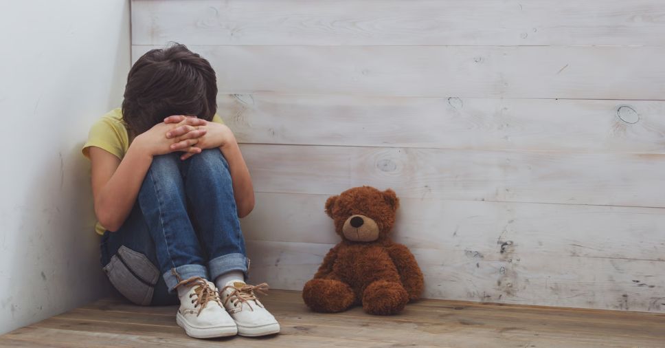 Criança encolhida no canto ao lado de urso de pelúcia retrata a ansiedade infantil autista.