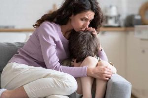 Entenda mais sobre suporte familiar e mães de autistas Mãe preocupada e carinhosa confortando o seu filho sentado no sofá, ilustrando as mães de autistas.
