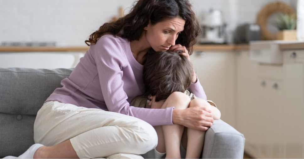 Mãe preocupada e carinhosa confortando o seu filho sentado no sofá, ilustrando as mães de autistas.